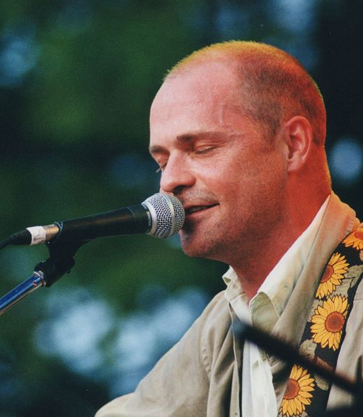 Gord Downie singing into a microphone with his eyes closed while wearing a biege dress shirt with a sunflower strap going over his shoulder.